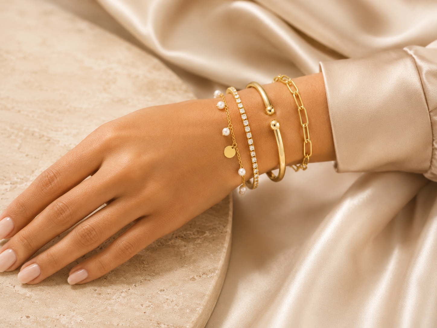 Gold-plated bracelet stack with chain, cuff, CZ and pearl charm styles on a warm neutral background